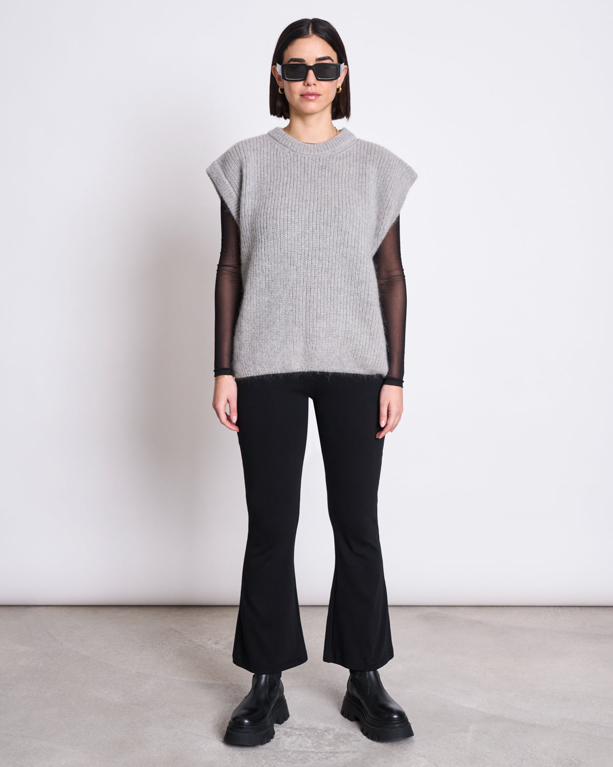 A person stands against a plain white wall wearing black sunglasses, the jan-n-june WOOLY SLIPOVER THANDA LIGHT GREY sweater over a black long-sleeve top, paired with black flared pants and chunky boots.