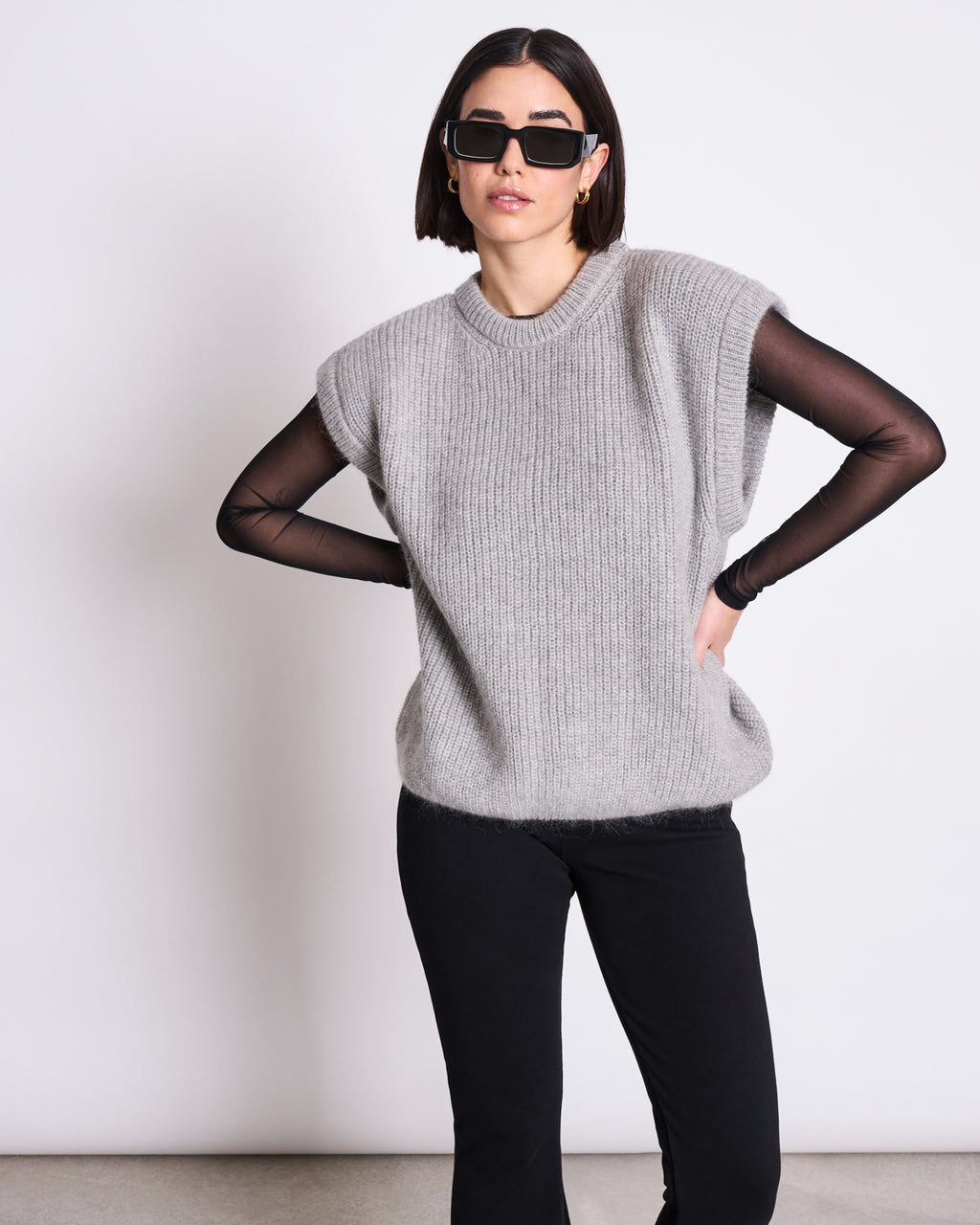 A woman wearing black sunglasses, the jan-n-june WOOLY SLIPOVER THANDA LIGHT GREY over a sheer black long-sleeve top, and black pants stands against a plain white background with one hand on her hip.