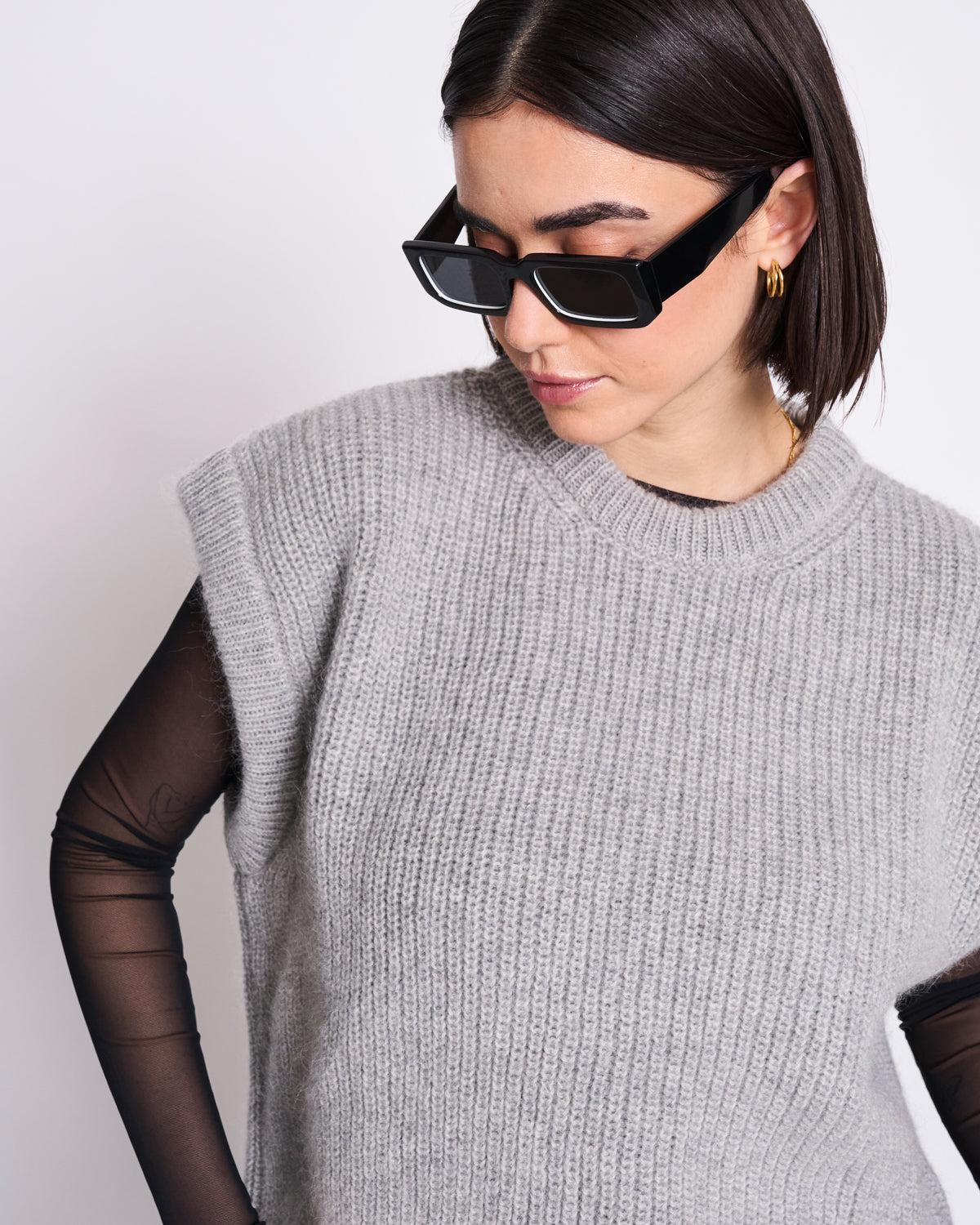 A woman with straight dark hair wears rectangular black sunglasses, gold hoop earrings, and the jan-n-june WOOLY SLIPOVER THANDA LIGHT GREY over a black sheer long-sleeve top, looking downward against a plain white background.