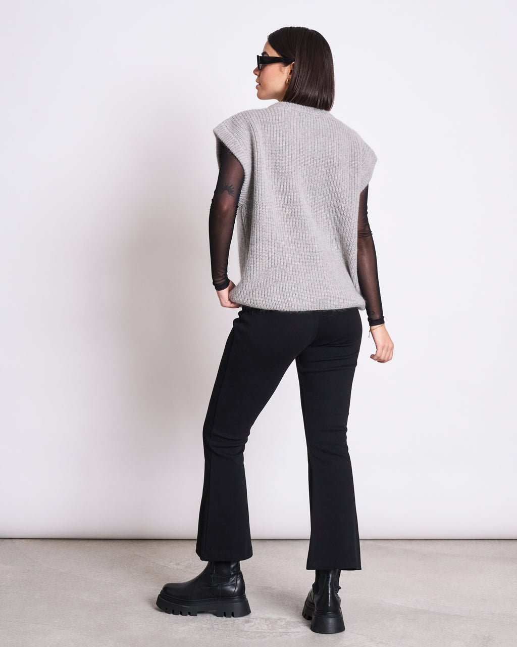 A woman stands with her back to the camera wearing jan-n-june’s WOOLY SLIPOVER THANDA LIGHT GREY over a black sheer long-sleeve top, paired with black pants, chunky black boots, and sunglasses against a white background.