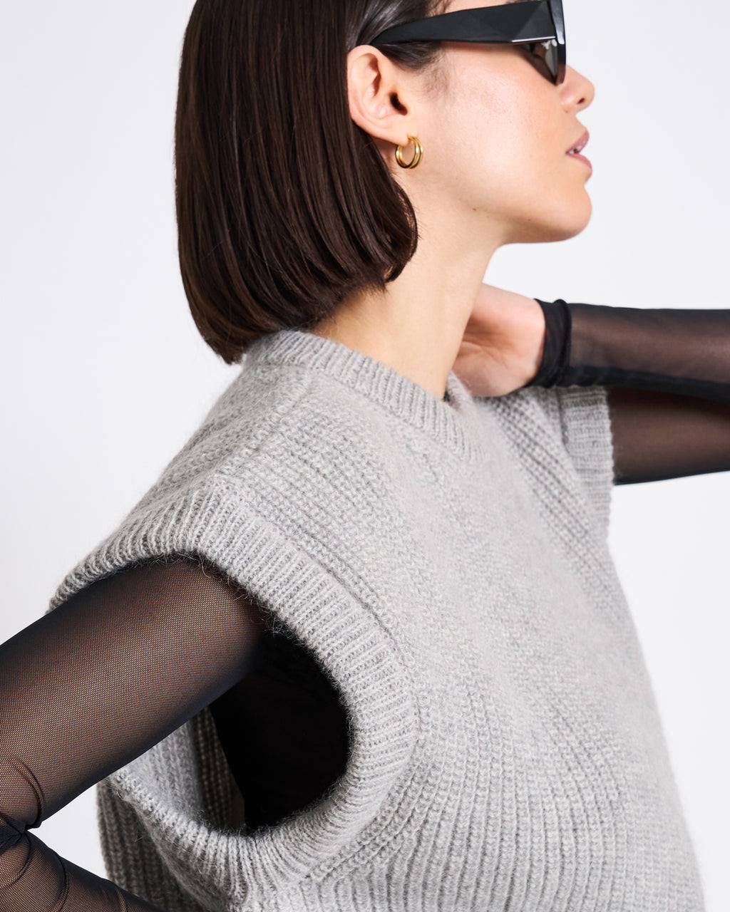 A woman with short dark hair wears black sunglasses, a gold hoop earring, a sheer black top, and the WOOLY SLIPOVER THANDA LIGHT GREY by jan-n-june in an oversized fit. She is turned to the side against a plain white background.