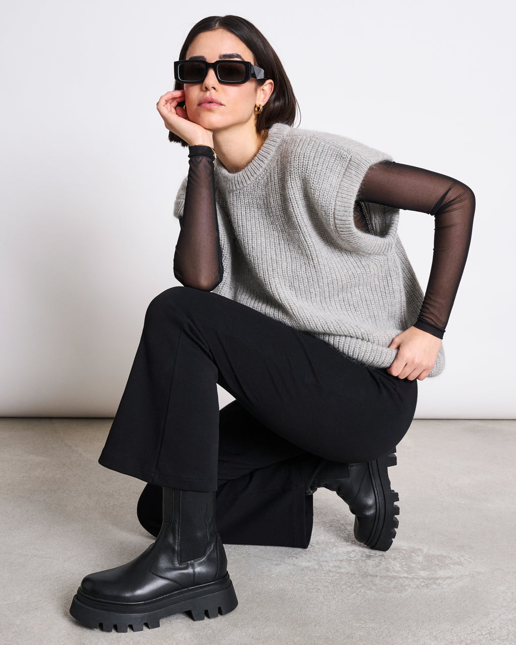 A woman in black flared pants, chunky boots, a sheer long-sleeve top, the jan-n-june WOOLY SLIPOVER THANDA LIGHT GREY vest, and sunglasses kneels on one knee against a plain background, posing with one hand on her chin.