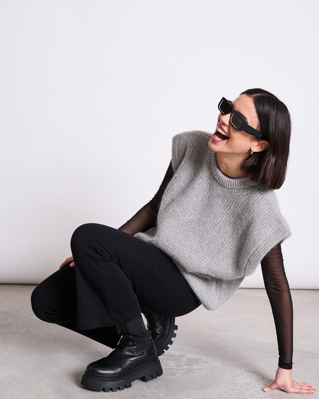 A woman with a bob haircut, wearing black sunglasses and the jan-n-june WOOLY SLIPOVER THANDA LIGHT GREY over a black top, black pants, and chunky boots, crouches and laughs against a plain white background.