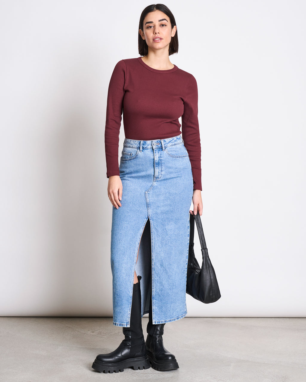 A woman stands against a white wall wearing the jan-n-june RIB LONGSLEEVE MURI INTENSE RUST GOTS, a light blue denim midi skirt with front slit, black chunky boots, and holding a black handbag—a true wardrobe essential.