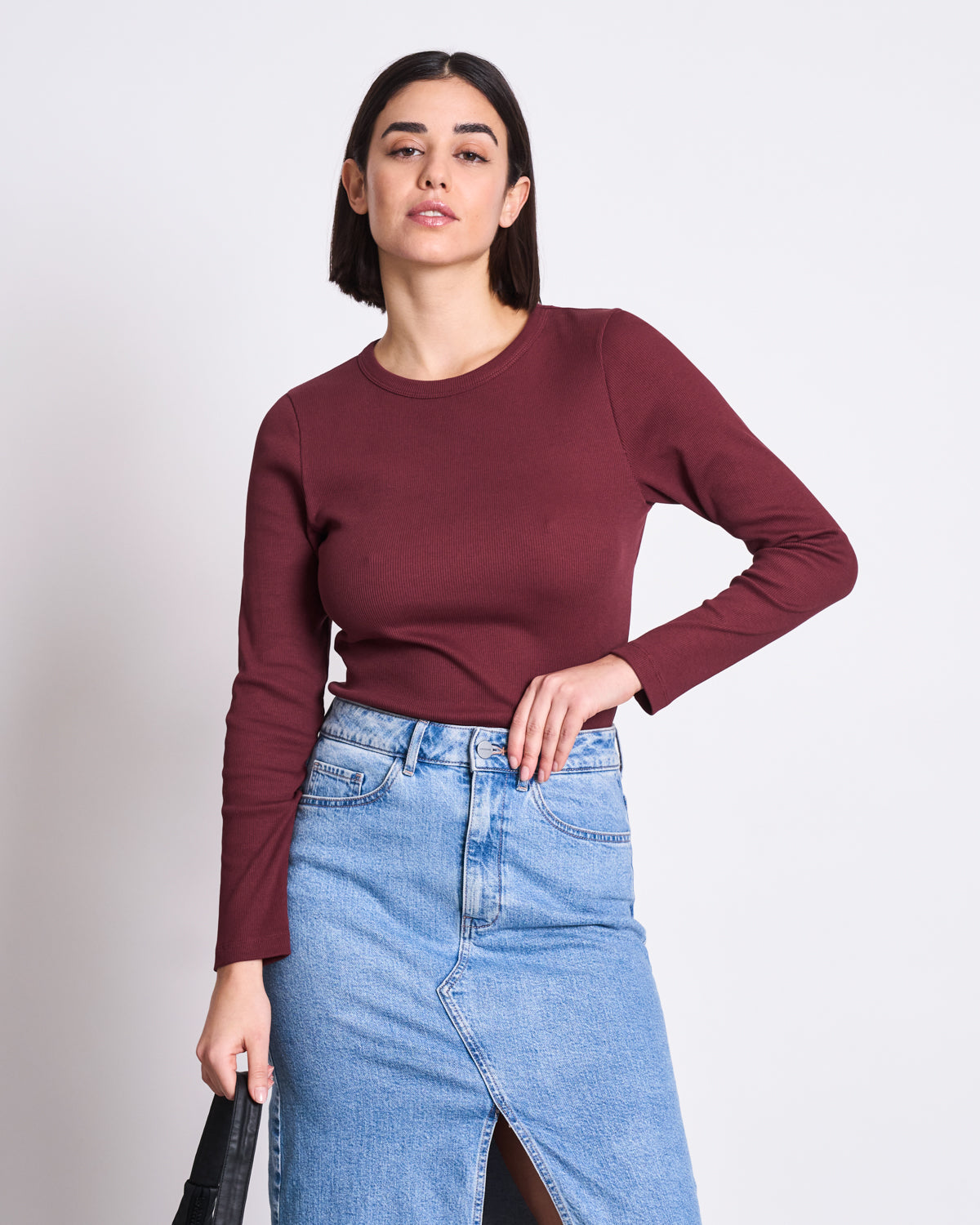 A woman with straight dark hair wears the jan-n-june RIB LONGSLEEVE MURI INTENSE RUST GOTS, paired with a high-waisted light blue denim skirt. She stands against a white background, holding a black bag—showcasing this versatile wardrobe essential.