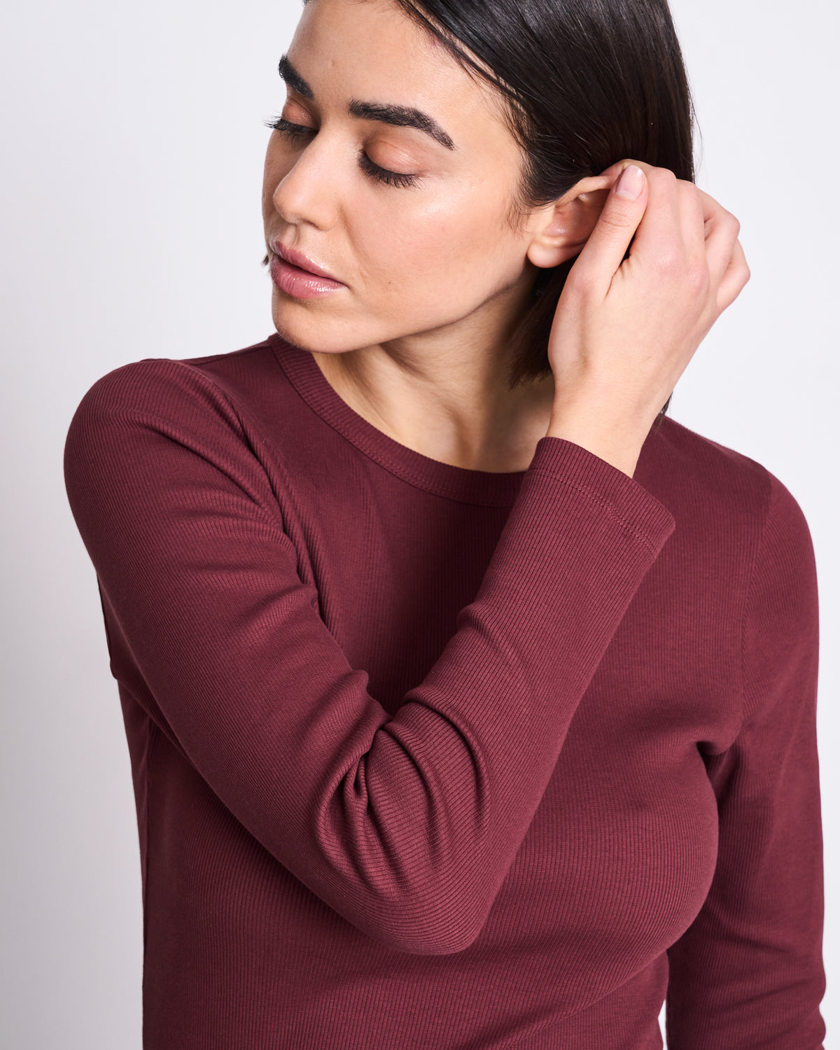 A woman with straight dark hair, wearing the jan-n-june RIB LONGSLEEVE MURI INTENSE RUST GOTS top, looks down and touches her ear with her right hand against a plain light background.