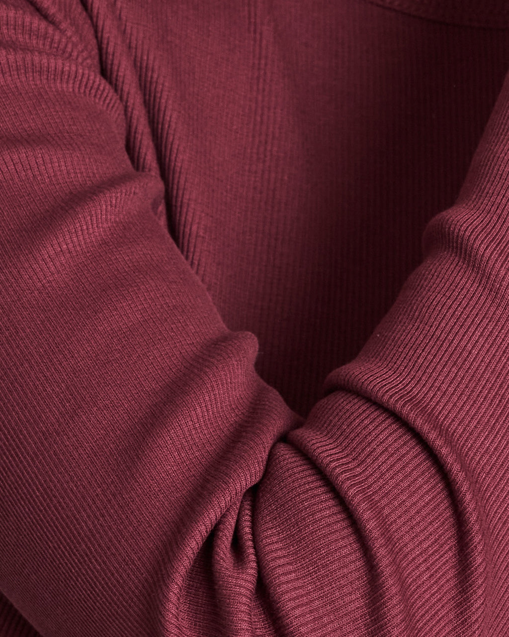 Close-up of an arm in the jan-n-june RIB LONGSLEEVE MURI INTENSE RUST GOTS—a slim, ribbed maroon shirt made from organic cotton, showing part of the torso and shoulder. A wardrobe essential for any collection.
