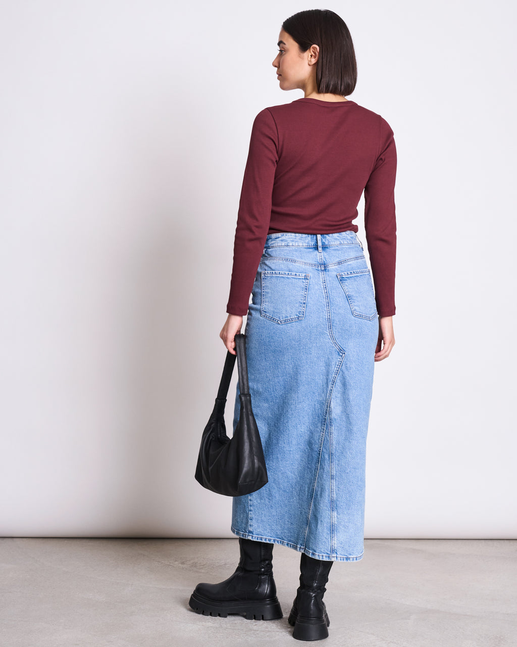 A person with short dark hair, wearing the jan-n-june RIB LONGSLEEVE MURI INTENSE RUST GOTS shirt, a light blue denim maxi skirt, black boots, and holding a black handbag stands facing away from the camera against a plain background.