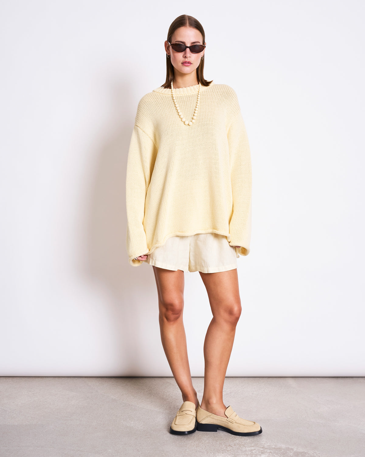 A woman with straight hair and a neutral expression stands against a white wall, wearing black sunglasses, cream shorts, beige loafers, a beaded necklace, and the jan-n-june JUMPER SONA BUTTERMILK GOTS oversized knit sweater.