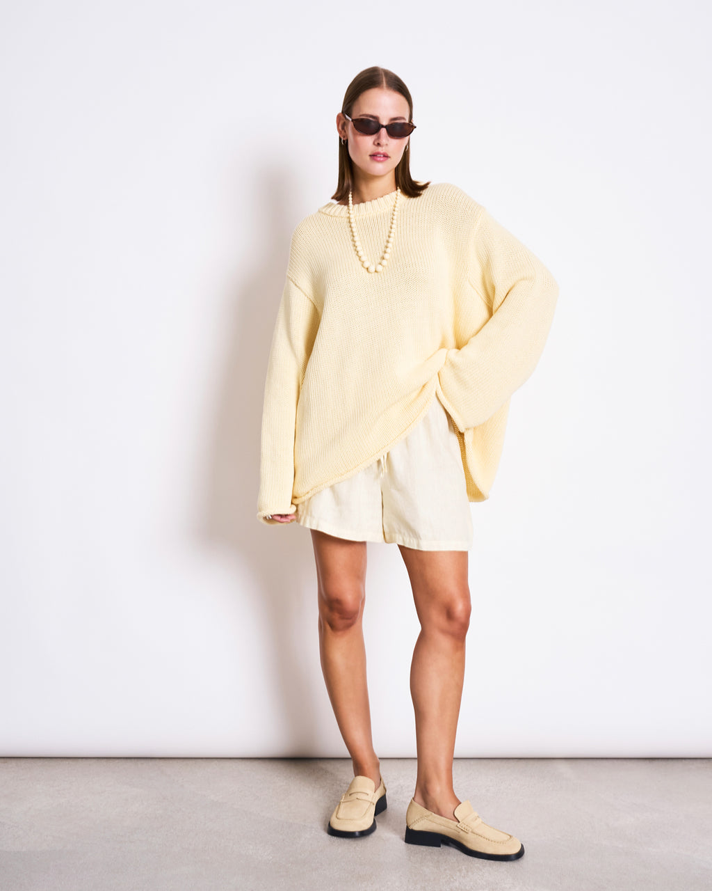 A woman wearing sunglasses, light shorts, a beaded necklace, beige loafers, and the jan-n-june JUMPER SONA BUTTERMILK GOTS cream sweater stands against a white wall with one hand on her hip.