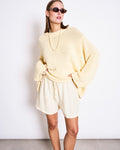 A woman poses against a white wall in the jan-n-june JUMPER SONA BUTTERMILK GOTS, an oversized pale yellow organic cotton sweater, styled with cat-eye sunglasses, a chunky necklace, and loose cream-colored shorts.