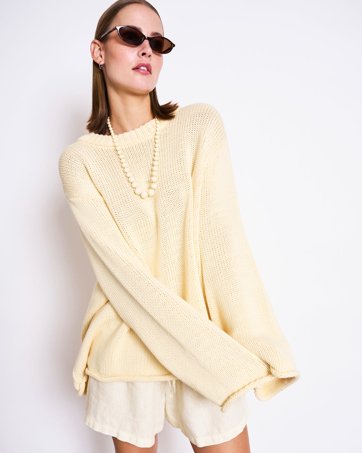 A woman in sunglasses wears the jan-n-june JUMPER SONA BUTTERMILK GOTS—an oversized, cream, organic cotton sweater—paired with cream shorts and a beaded necklace, posing confidently against a plain white background.