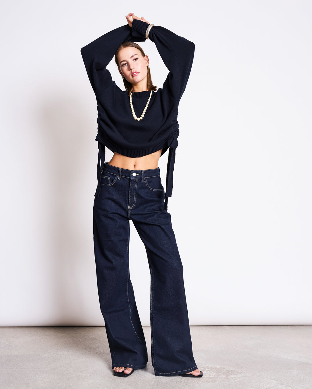 A woman poses confidently against a white background in jan-n-june’s WIDE LEG LOWRISE JEANS TACNA DARK BLUE DENIM GOTS, paired with a cropped black sweater, black sandals, and a chunky necklace. Her arms are raised above her head.