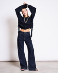 A woman poses confidently against a white background in jan-n-june’s WIDE LEG LOWRISE JEANS TACNA DARK BLUE DENIM GOTS, paired with a cropped black sweater, black sandals, and a chunky necklace. Her arms are raised above her head.
