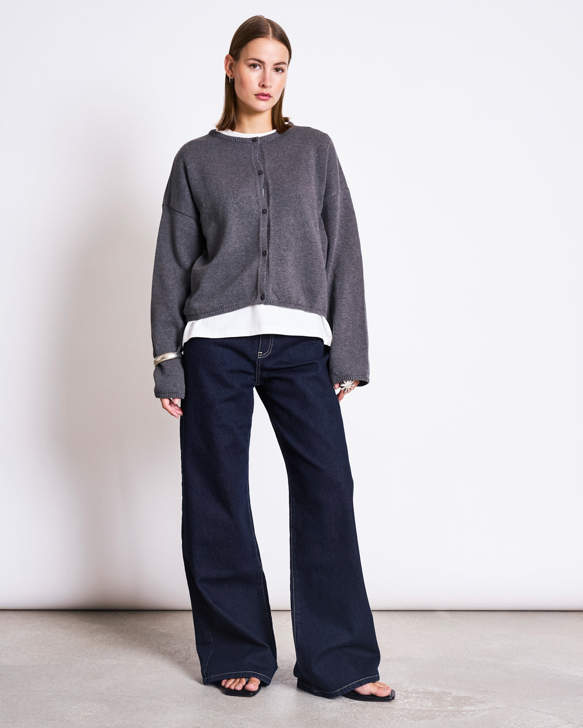 A woman stands against a white wall wearing the jan-n-june LIGHT CARDIGAN ASAS DARK GREY GOTS over a white shirt, paired with dark wide-leg jeans and black sandals. She looks relaxed, her hands at her sides.