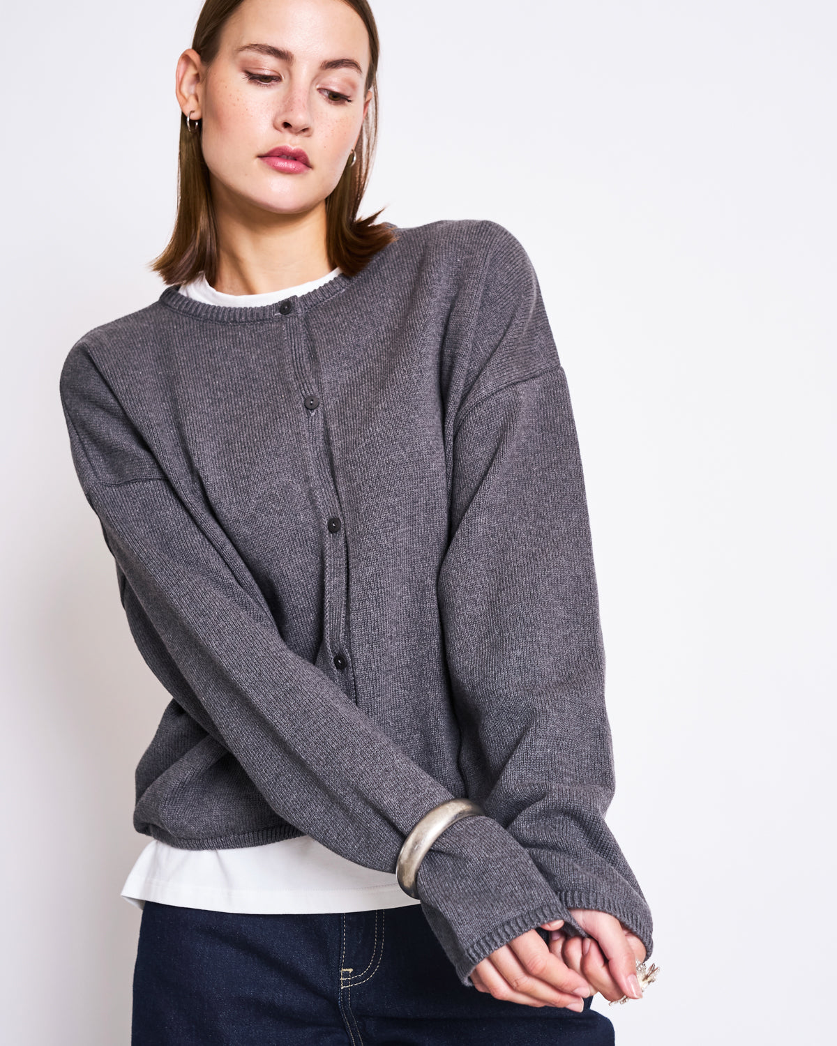 A young woman with straight brown hair wears the jan-n-june LIGHT CARDIGAN ASAS DARK GREY GOTS, made from organic cotton, over a white shirt. She stands against a plain white background, arms relaxed and head slightly tilted down.