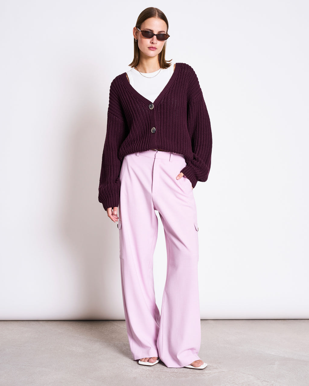 A woman stands against a plain white wall, one hand in her pocket, wearing jan-n-june’s CHUNKY CARDIGAN LENA CHERRY LACQUER GOTS over a white shirt, with sunglasses and loose light pink pants.