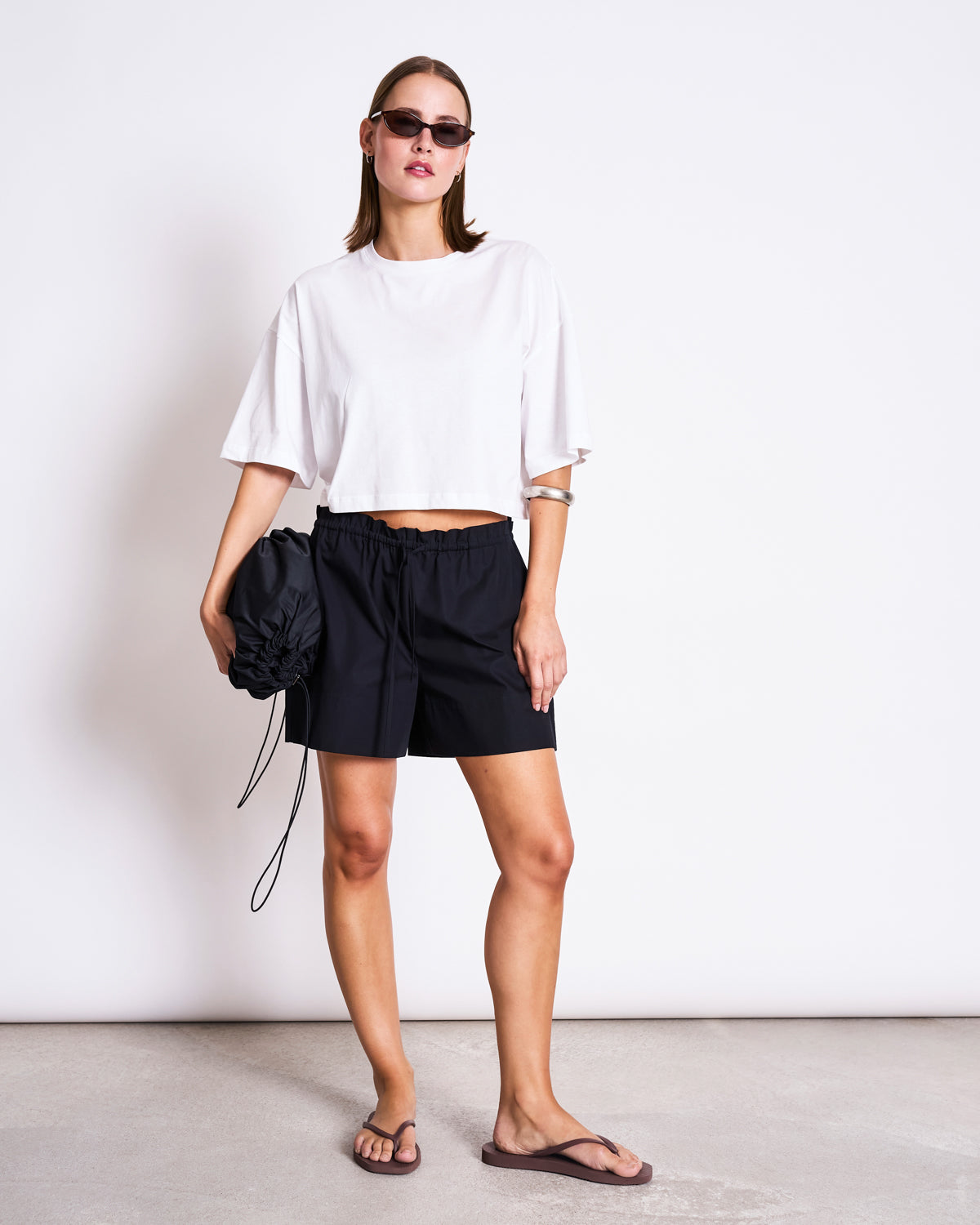 A woman stands against a white wall wearing sunglasses, black shorts, flip-flops, holding a black ruched bag, and the jan-n-june CROPPED BOXY SHIRT LUPA WHITE with an oversized fit.