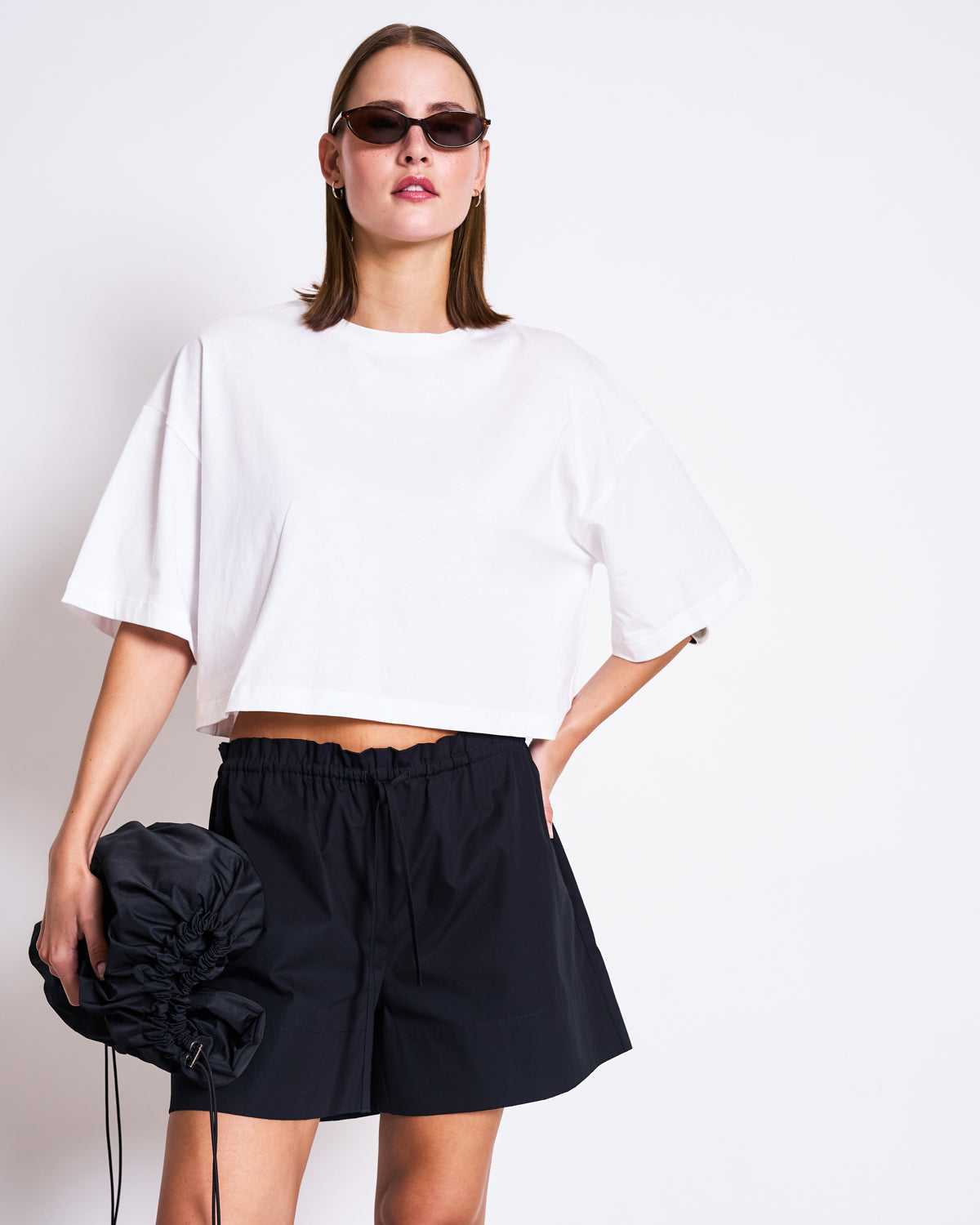 A woman wears the jan-n-june CROPPED BOXY SHIRT LUPA WHITE, styled with black high-waisted shorts, sunglasses, and a black ruched handbag, standing against a white background.