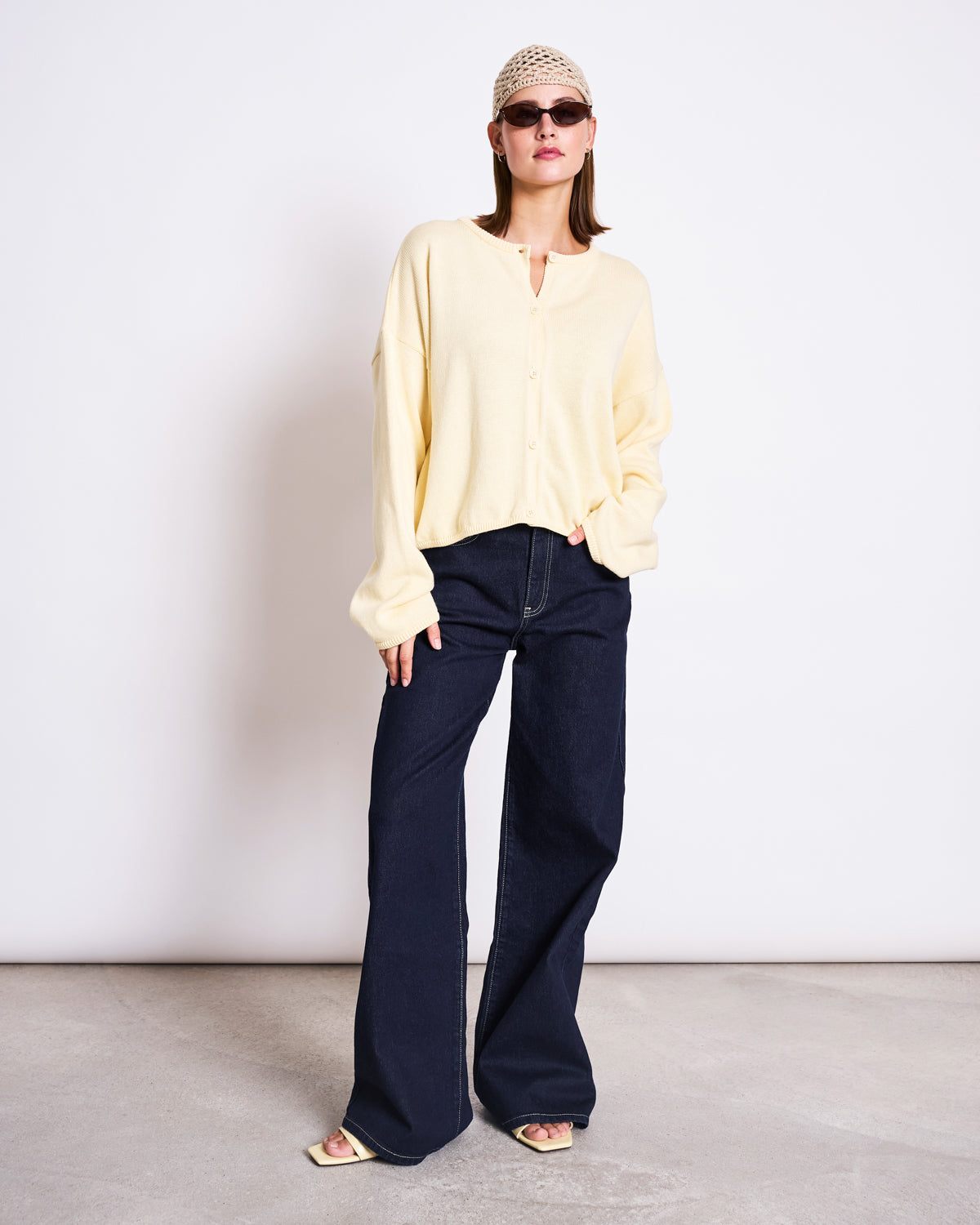 Wearing sunglasses, a beige head covering, the jan-n-june LIGHT CARDIGAN ASAS BUTTERMILK GOTS, wide-leg dark blue jeans, and open-toed sandals, this person stands against a plain light background—embodying sustainable style.