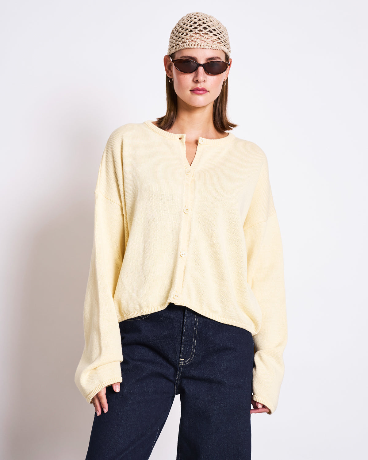A woman in the jan-n-june LIGHT CARDIGAN ASAS BUTTERMILK GOTS, made from organic cotton, pairs it with dark jeans, sunglasses, and a crocheted beanie, embracing sustainable style against a plain white backdrop.
