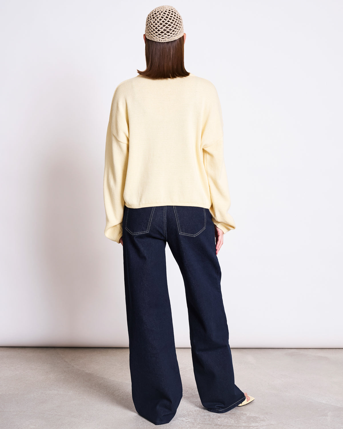 A person stands facing away in jan-n-junes LIGHT CARDIGAN ASAS BUTTERMILK GOTS, paired with wide-leg dark blue jeans, a cream crochet beanie, and light shoes on a plain concrete floor for an effortless sustainable look.