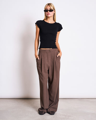 A woman wearing sunglasses, the jan-n-june MINIRIB T-SHIRT REN BLACK GOTS with a cropped fit, loose brown trousers, and black textured shoes stands with hands in her pockets against a plain white background.