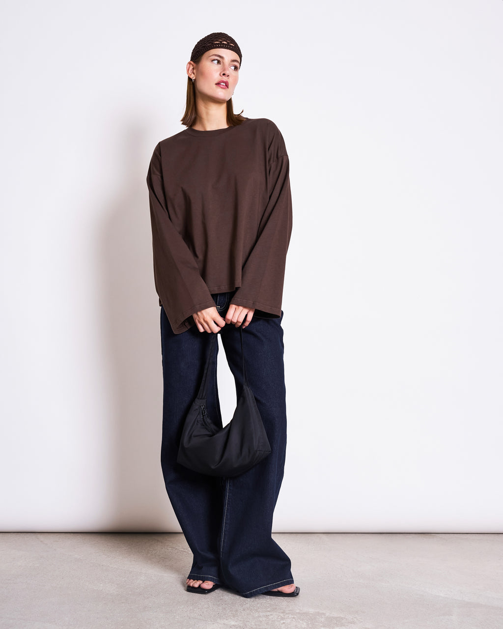 A woman stands against a white wall wearing the jan-n-june BOXY LONGSLEEVE ANKIRA MUD, paired with wide-leg dark jeans, black sandals, a patterned headband, and holding a black bag in front of her.