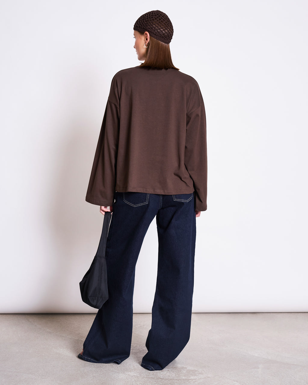 A person stands with their back to the camera, wearing jan-n-junes BOXY LONGSLEEVE ANKIRA MUD shirt, wide-leg dark jeans, a brown mesh beanie, and holding a small black bag against a plain light background.