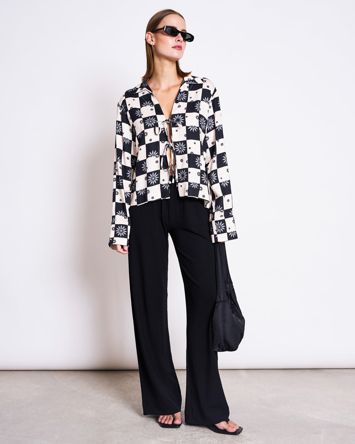 Wearing sunglasses, loose black pants, and flip-flops, a person stands against a white wall holding a black bag—showcasing effortless style in the jan-n-june TIE BLOUSE KALUZ FLORAL SQUARE PRINT.