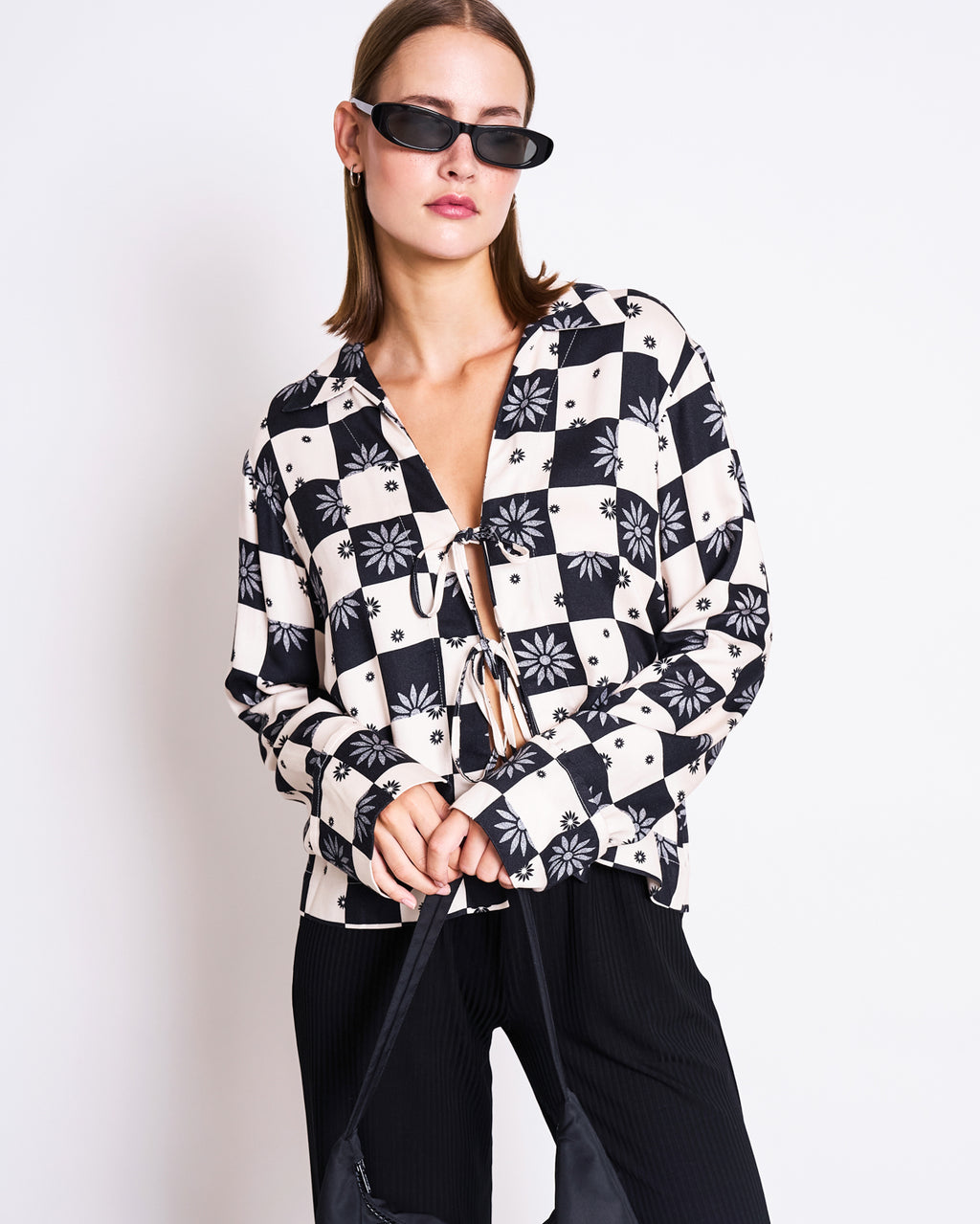 A woman stands against a plain white background wearing black sunglasses, black pants, and the jan-n-june TIE BLOUSE KALUZ FLORAL SQUARE PRINT with front ties, holding a black bag—a chic nod to sustainable fashion.