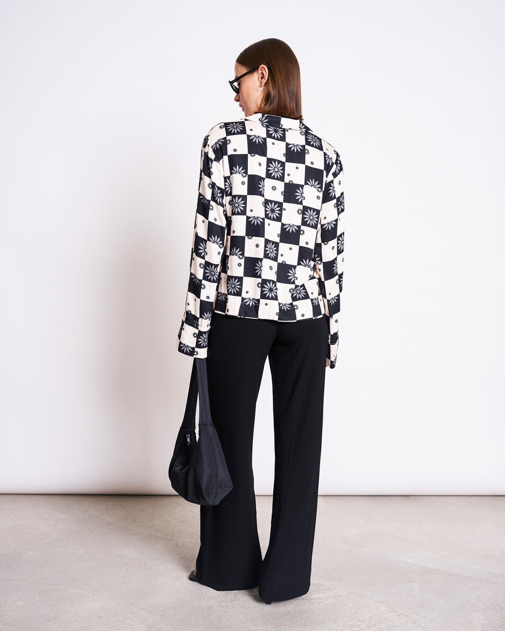 A person wearing sunglasses, a checkered jacket with sun designs over the jan-n-june TIE BLOUSE KALUZ FLORAL SQUARE PRINT, and black wide-leg pants stands facing away, holding a black bag in their left hand against a plain white wall.