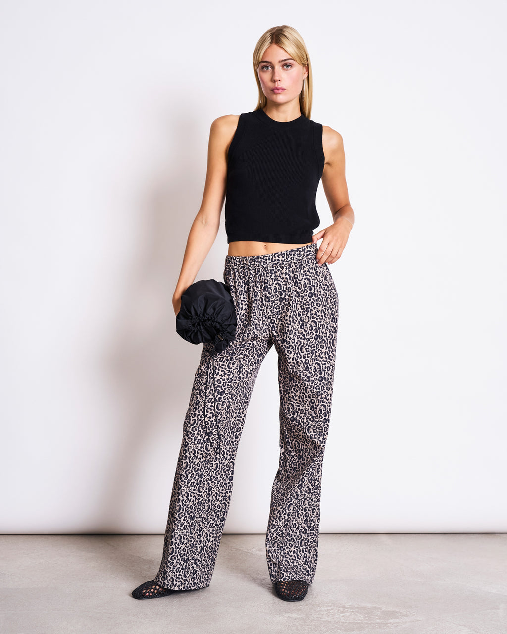 A woman stands against a white wall, wearing jan-n-june’s high-waisted organic cotton pants in the SOBY LEO print with a sleeveless black crop top. She holds a black scrunchie bag and looks forward with a neutral expression.