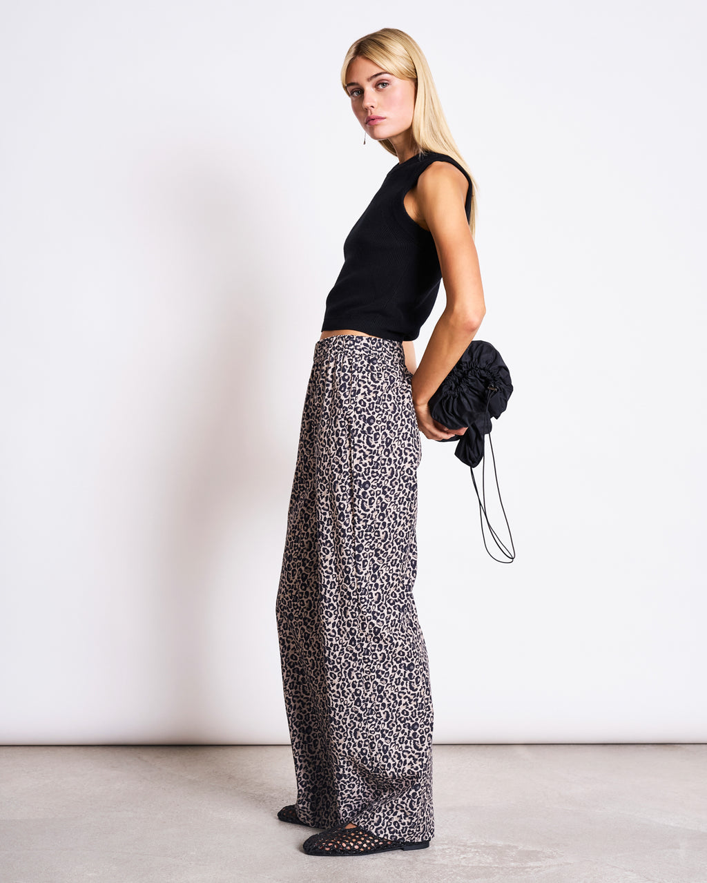 A woman with long blonde hair stands sideways in a minimal studio, wearing jan-n-junes COTTON PANTS SOBY LEO PRINT—loose-fitting organic cotton leopard print pants—with a sleeveless black top and holding a scrunched black bag behind her.