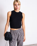 A woman with blonde hair models jan-n-june’s COTTON PANTS SOBY LEO PRINT, sleeveless black top, and holds a black drawstring bag, standing against a plain white background.