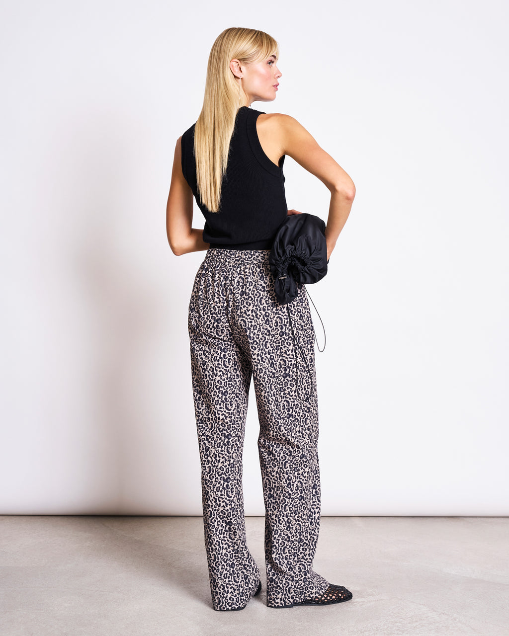 A woman with straight blonde hair, facing away, wears jan-n-junes COTTON PANTS SOBY LEO PRINT—loose, high-waisted leopard-print cotton pants—paired with a sleeveless black top and holds a black bag against a plain white background.