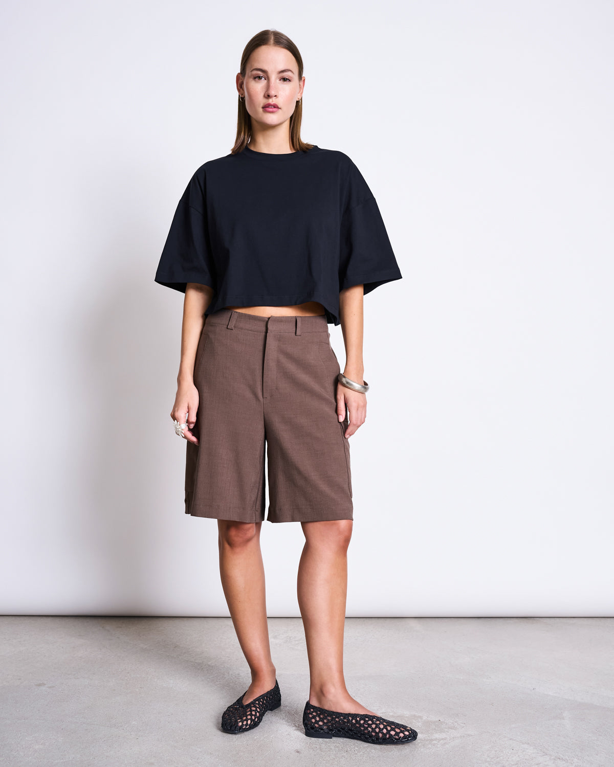 A woman with straight hair and a neutral expression stands against a white wall, wearing the jan-n-june CROPPED BOXY SHIRT LUPA BLACK, knee-length brown shorts, and black woven slip-on shoes.