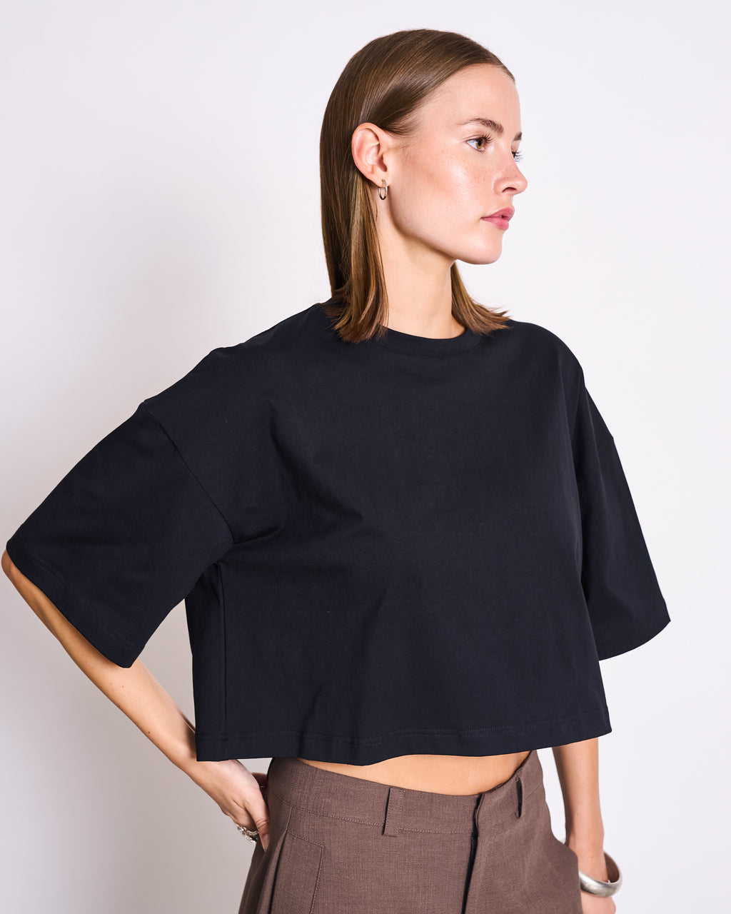 A woman with straight brown hair models the jan-n-june CROPPED BOXY SHIRT LUPA BLACK, styled oversized and cropped. She stands with one hand on her hip, gazing to the side against a plain white background.
