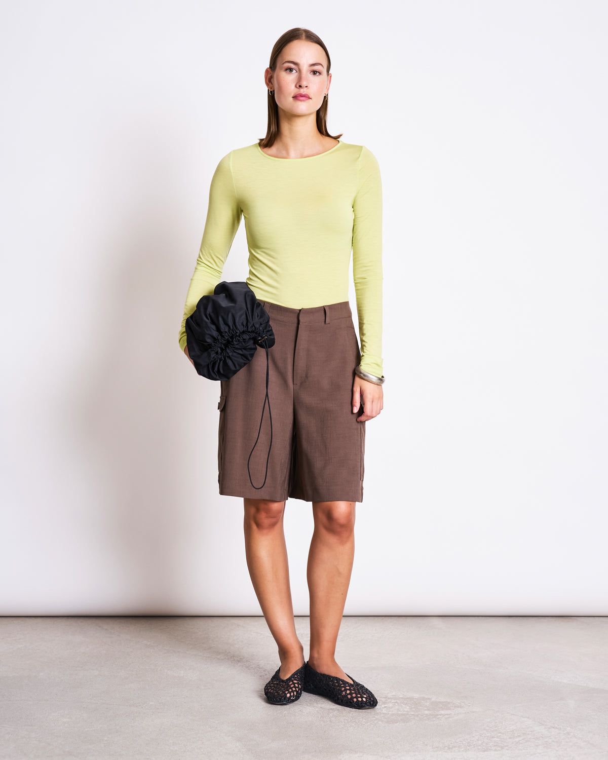A woman stands against a white wall wearing the jan-n-june TENCEL™ LONGSLEEVE CIVA GRANNY SMITH top, brown knee-length shorts, black slip-on shoes, and holding a black scrunched bag.