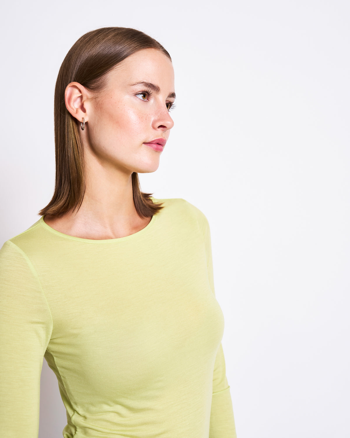 A woman with straight brown hair, wearing the jan-n-june TENCEL™ LONGSLEEVE CIVA GRANNY SMITH, stands against a plain white background and gazes to the side with a neutral expression.