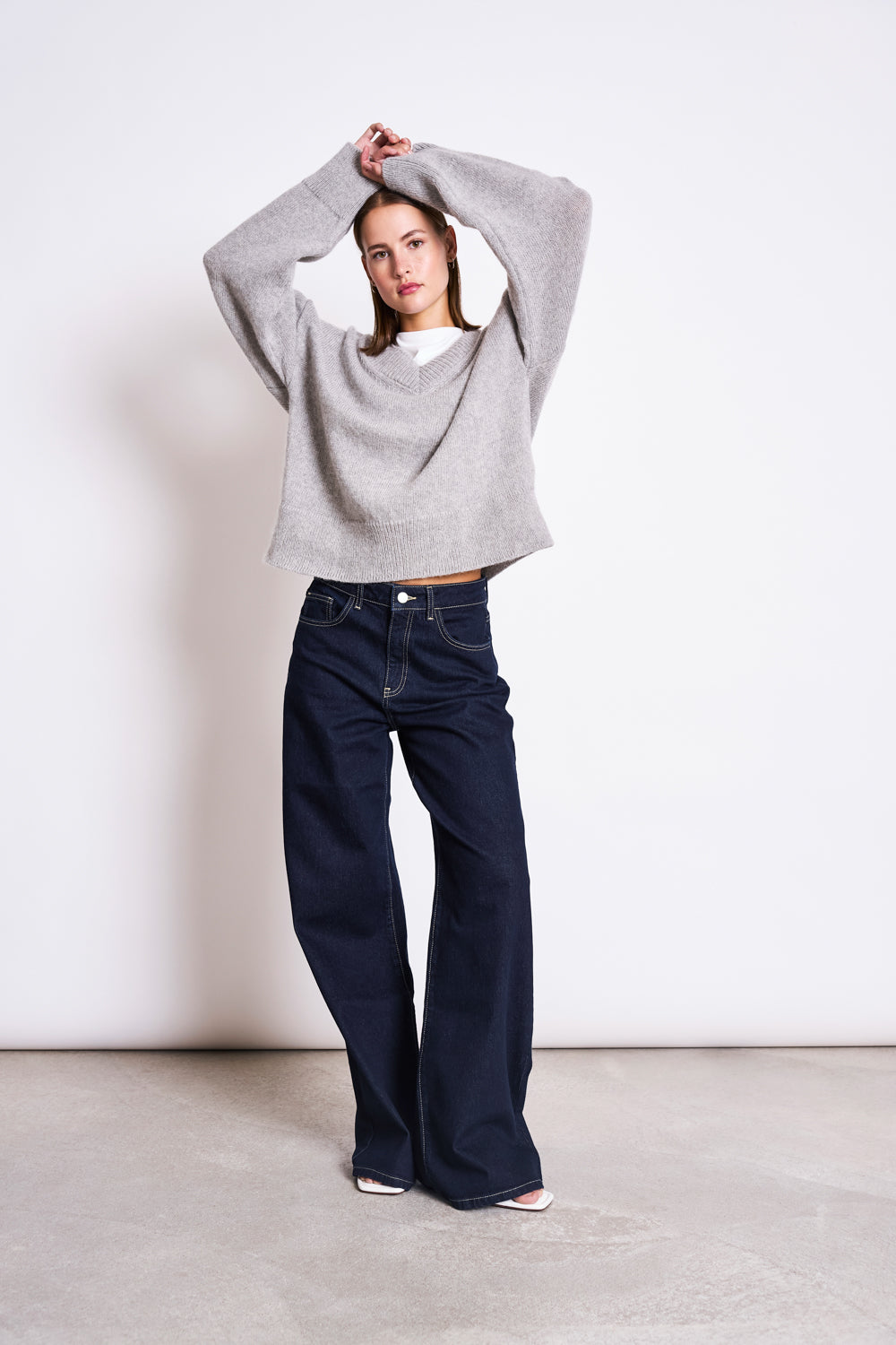 A woman poses indoors against a plain white wall, confidently wearing the jan-n-june REC. MOHAIR V-NECK JUMPER YICA in light grey over a white top and wide-leg dark blue jeans, with her arms raised and crossed above her head.