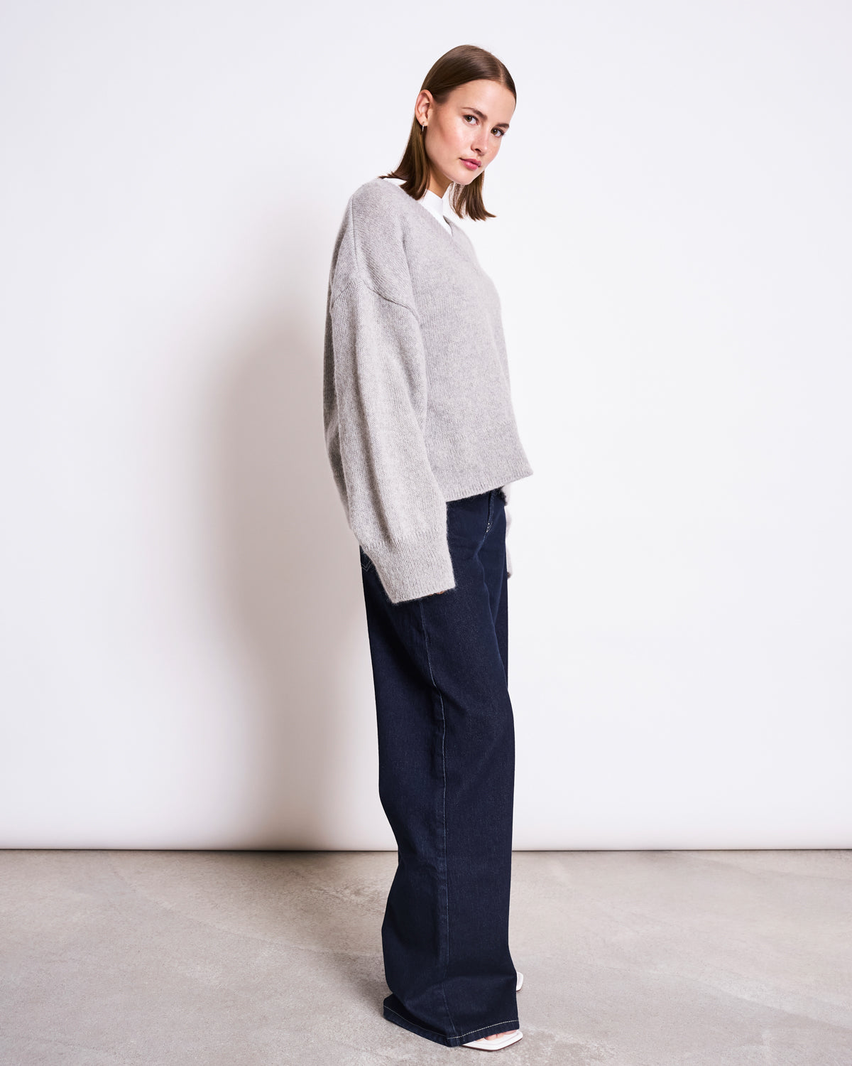 A woman with straight, shoulder-length brown hair and a neutral expression stands sideways against a plain white background, wearing the jan-n-june REC. MOHAIR V-NECK JUMPER YICA LIGHT GREY, wide-leg dark jeans, and white shoes.