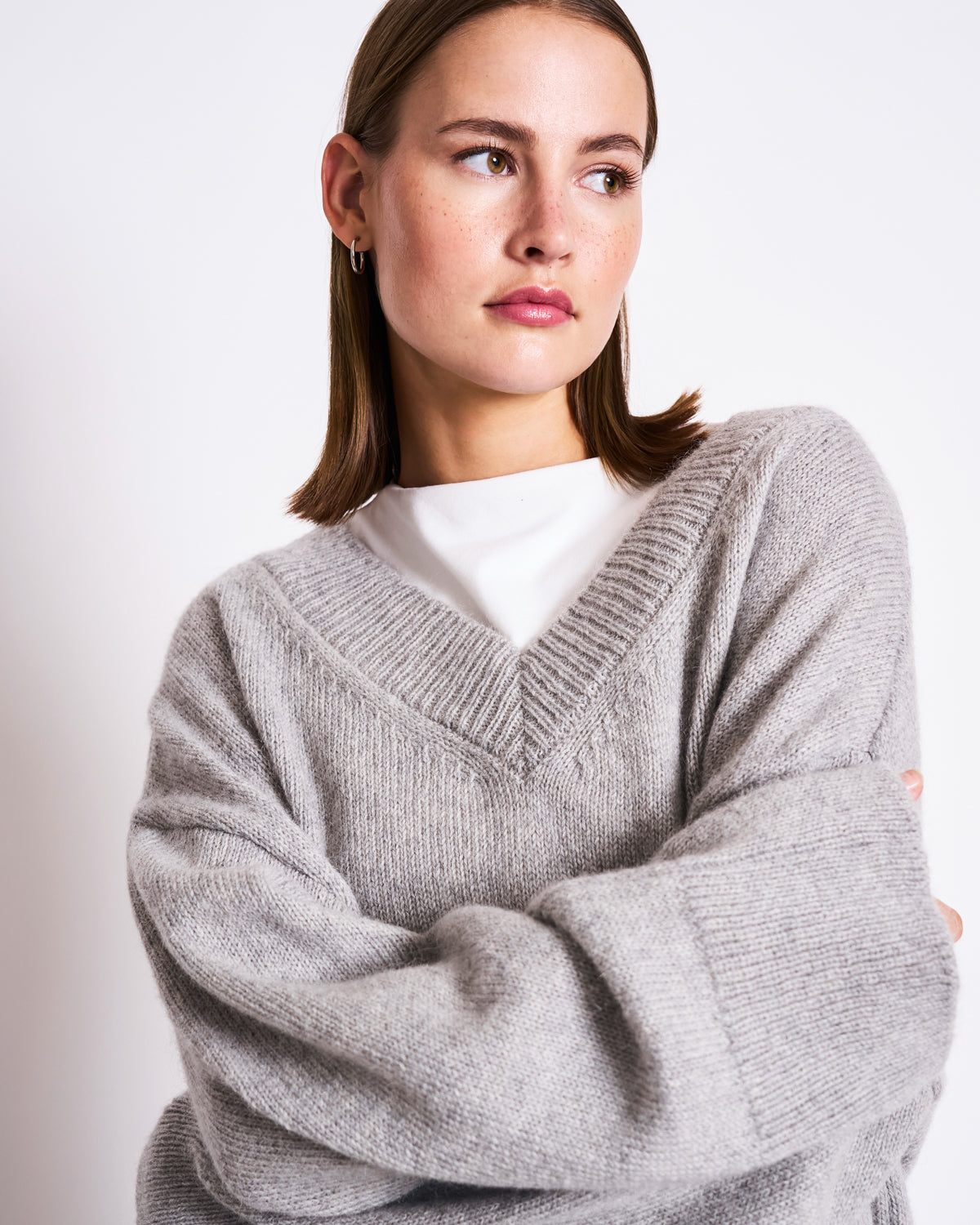 A woman with straight brown hair wears the jan-n-june REC. MOHAIR V-NECK JUMPER YICA in light grey, styled loosely over a white top, gazing to the side with arms crossed against a plain light background.