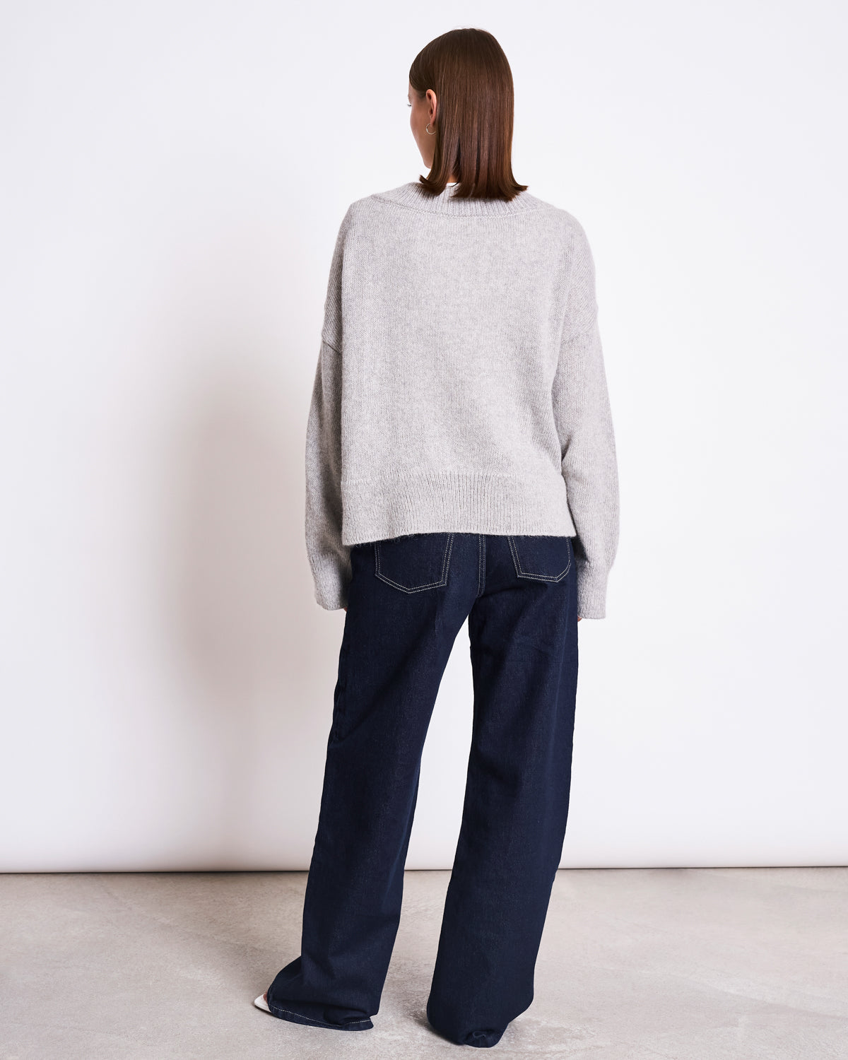 A person with straight brown hair, wearing the jan-n-june REC. MOHAIR V-NECK JUMPER YICA LIGHT GREY and dark blue wide-leg jeans, stands facing away from the camera against a plain white wall.