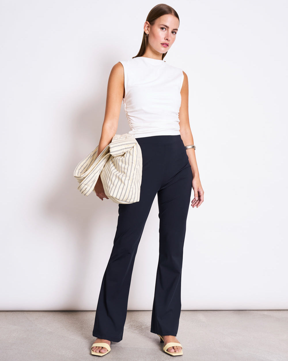 A woman with straight hair and a neutral expression poses against a white background in jan-n-junes ECONYL® FLARED LEGGINGS NORE BLACK, a sleeveless white top, light sandals, and holds a striped jacket over one arm.