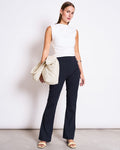 A woman with straight hair and a neutral expression poses against a white background in jan-n-junes ECONYL® FLARED LEGGINGS NORE BLACK, a sleeveless white top, light sandals, and holds a striped jacket over one arm.