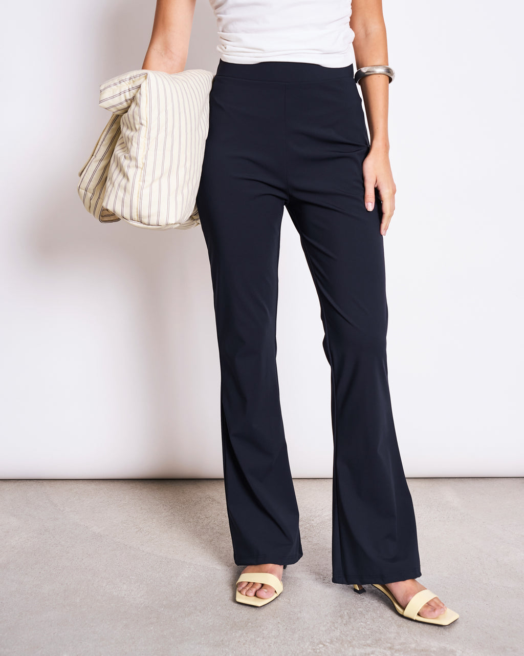 Wearing jan-n-june’s ECONYL® FLARED LEGGINGS NORE BLACK, a white top, and yellow open-toe heels, a person holds a striped tote bag and sports a chunky silver bracelet against a plain background.