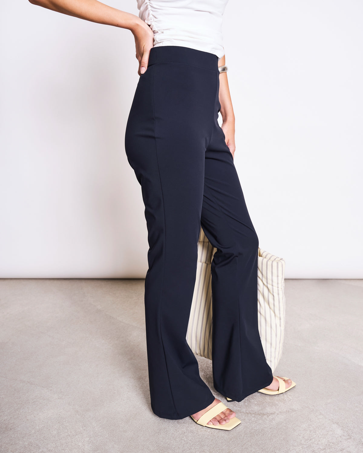 Someone wearing jan-n-june ECONYL® FLARED LEGGINGS NORE BLACK, a white top, and beige sandals stands on a light gray floor, holding a striped bag. The persons face is not visible.