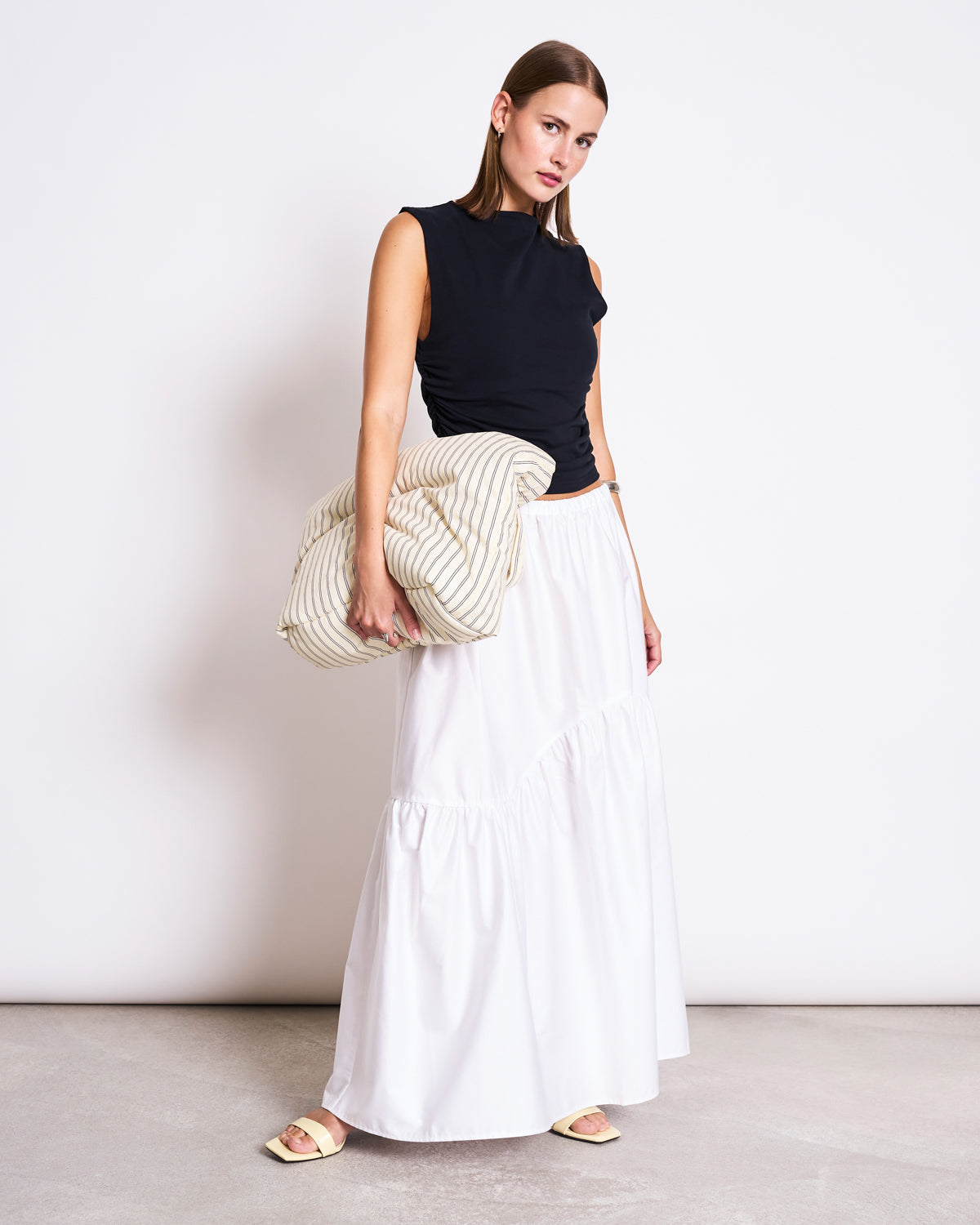 A woman stands against a plain white wall, wearing a sleeveless black top, the jan-n-june TIERED MAXI SKIRT ALCA WHITE, beige sandals, and holding a large striped cream clutch under her arm.