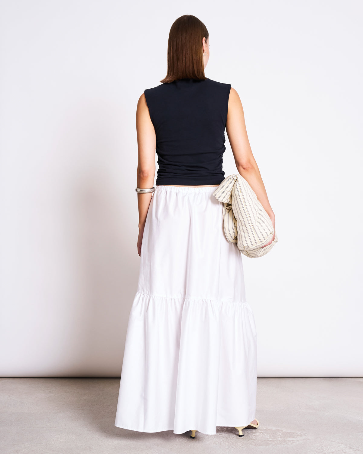 A woman stands facing away, wearing the jan-n-june TIERED MAXI SKIRT ALCA WHITE with a sleeveless black top, holding a striped bag on her left arm and light-colored heeled shoes, against a plain background.